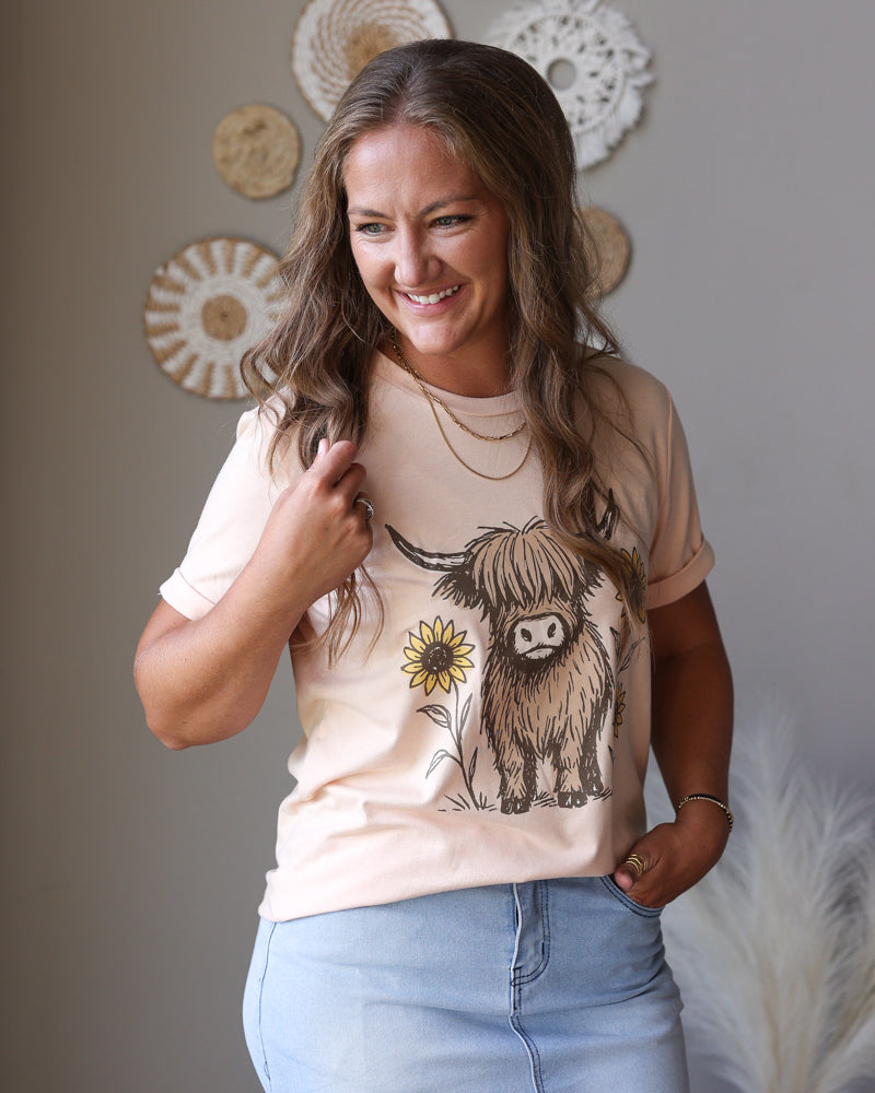 A woman smiling while wearing a light beige graphic t-shirt with a Highland cow and sunflower design, paired with light blue jean skirt, in a styled indoor setting with boho wall decor.