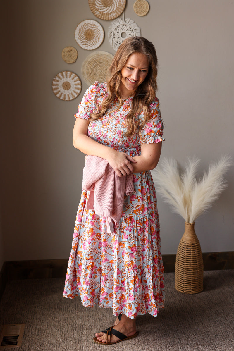 Woman in a floral dress holding a pink towel in a room with decorative wall art and a vase.