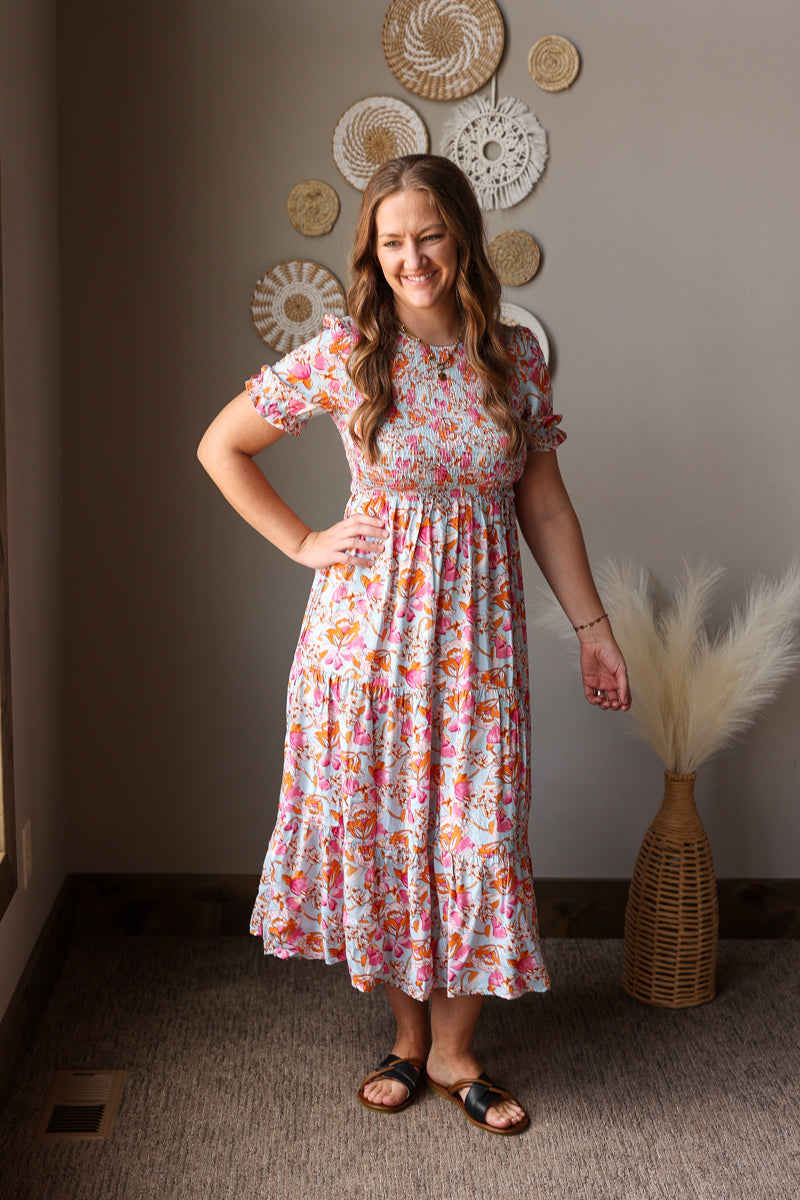 Woman in a floral dress standing in a room with decorative wall art and a vase.