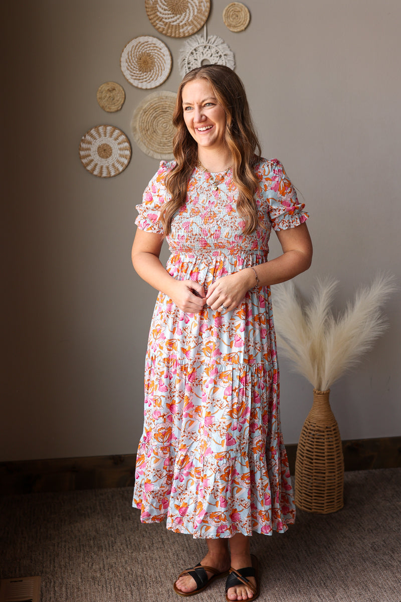 Woman in a floral dress standing in a room with decorative wall art and plants.