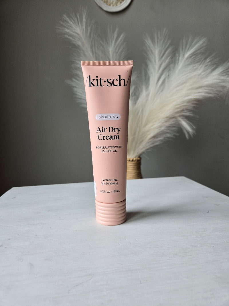 A pink tube of kit·sch Air Dry Cream smoothing air dry shampoo, styled diagonally on a white surface with a white feather prop against a dark gray background.