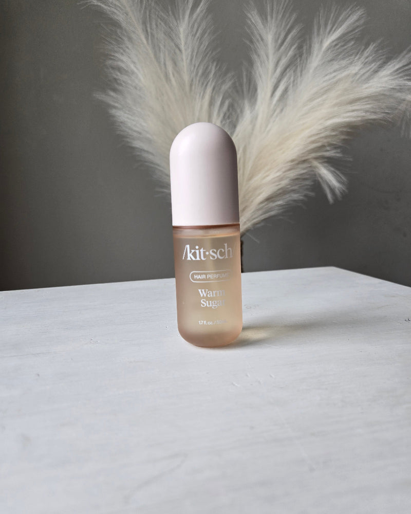 A bottle of Warm Sugar Kitsch Hair Perfume Body Mist Spray on a white surface, with a fluffy white decorative feather placed behind it against a dark gray background.
