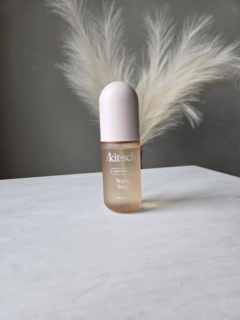 A bottle of Warm Sugar Kitsch Hair Perfume Body Mist Spray on a white surface, with a fluffy white decorative feather placed behind it against a dark gray background.
