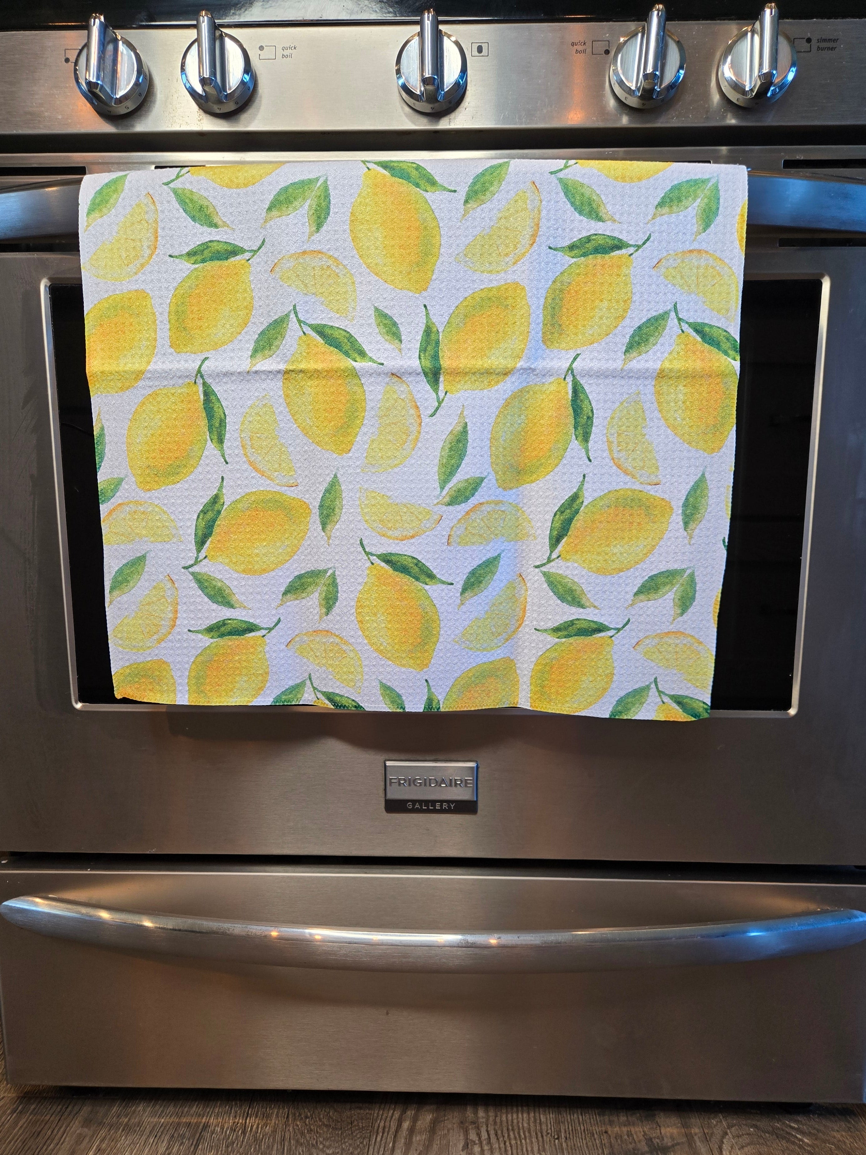 A white kitchen towel with a yellow lemon and green leaf pattern hanging on the handle of a stainless steel KitchenAid range.