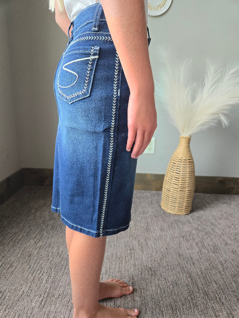 A person wearing a blue denim skirt with white contrast stitching, standing on a gray carpeted floor next to a woven decorative vase.