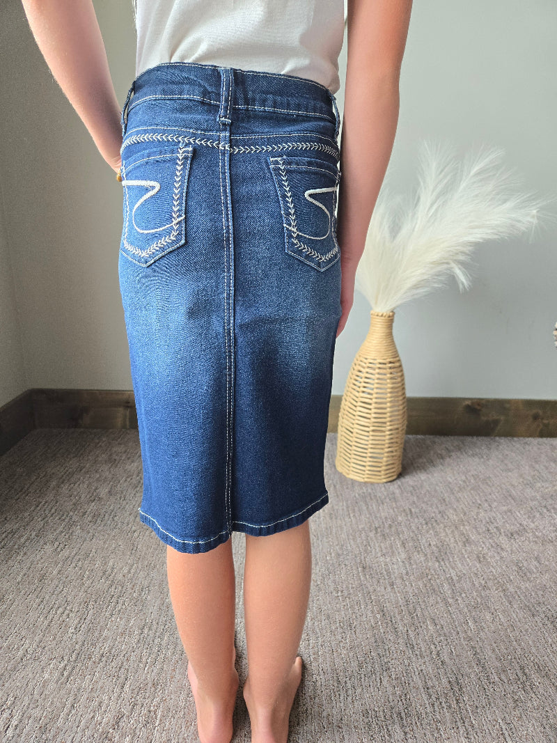 A person shown from the back wearing a blue denim pencil skirt with white contrast stitching and decorative stitching on the back pockets, standing on a gray carpeted floor next to a woven vase with pampas grass.