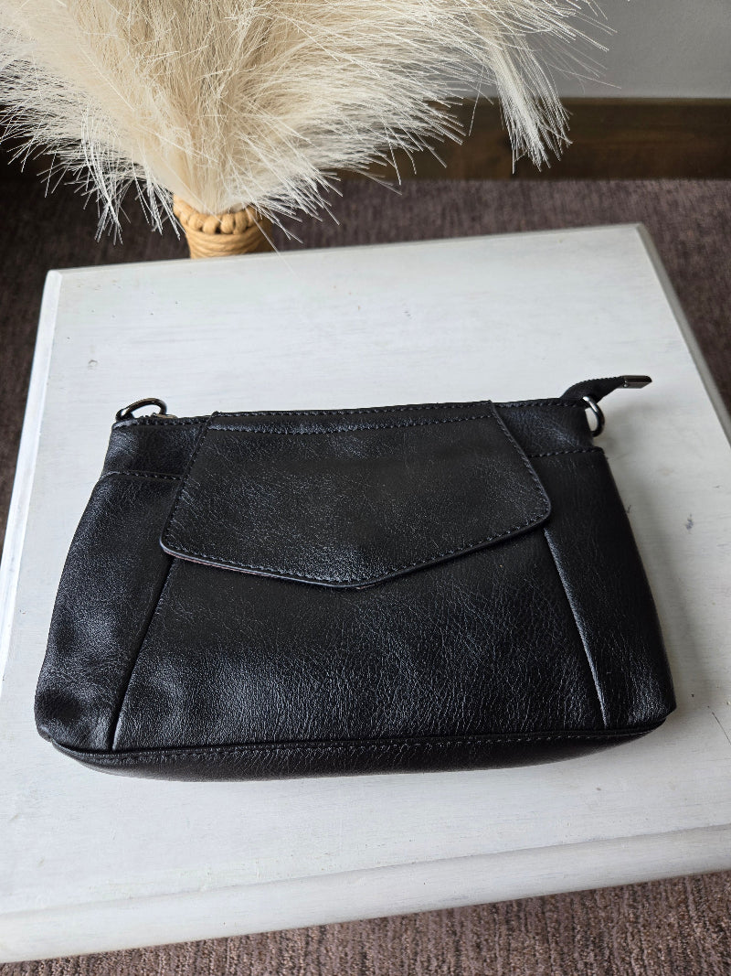 A black leather clutch or wristlet pouch with a front flap pocket, resting on a white wooden board with dried pampas grass in the background.