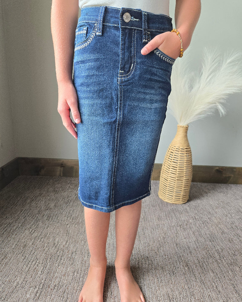 A person wearing a blue denim pencil skirt with contrast stitching, standing barefoot on a gray carpeted floor next to a woven vase with white pampas grass.