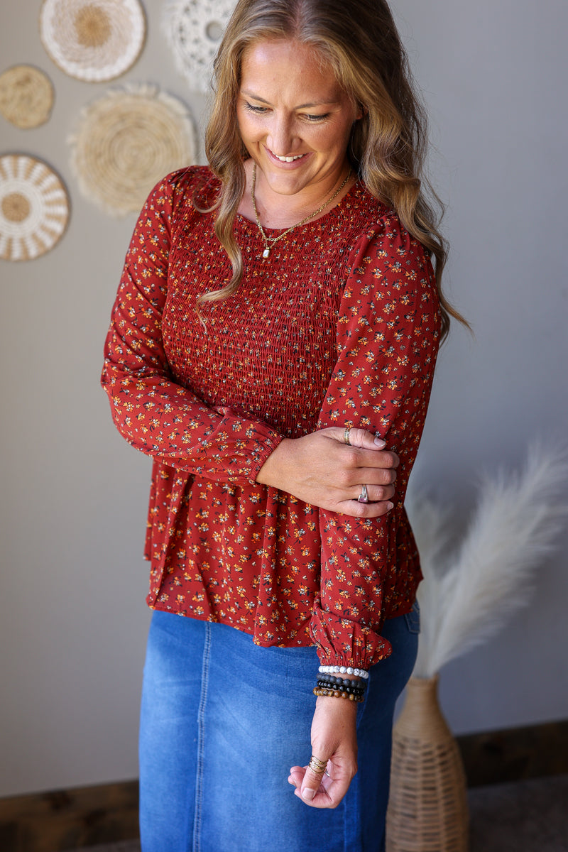 A woman wearing a rust red long-sleeve blouse with a small white floral pattern and blue jean skirt, standing in a room with neutral-colored walls and decorative wall art.