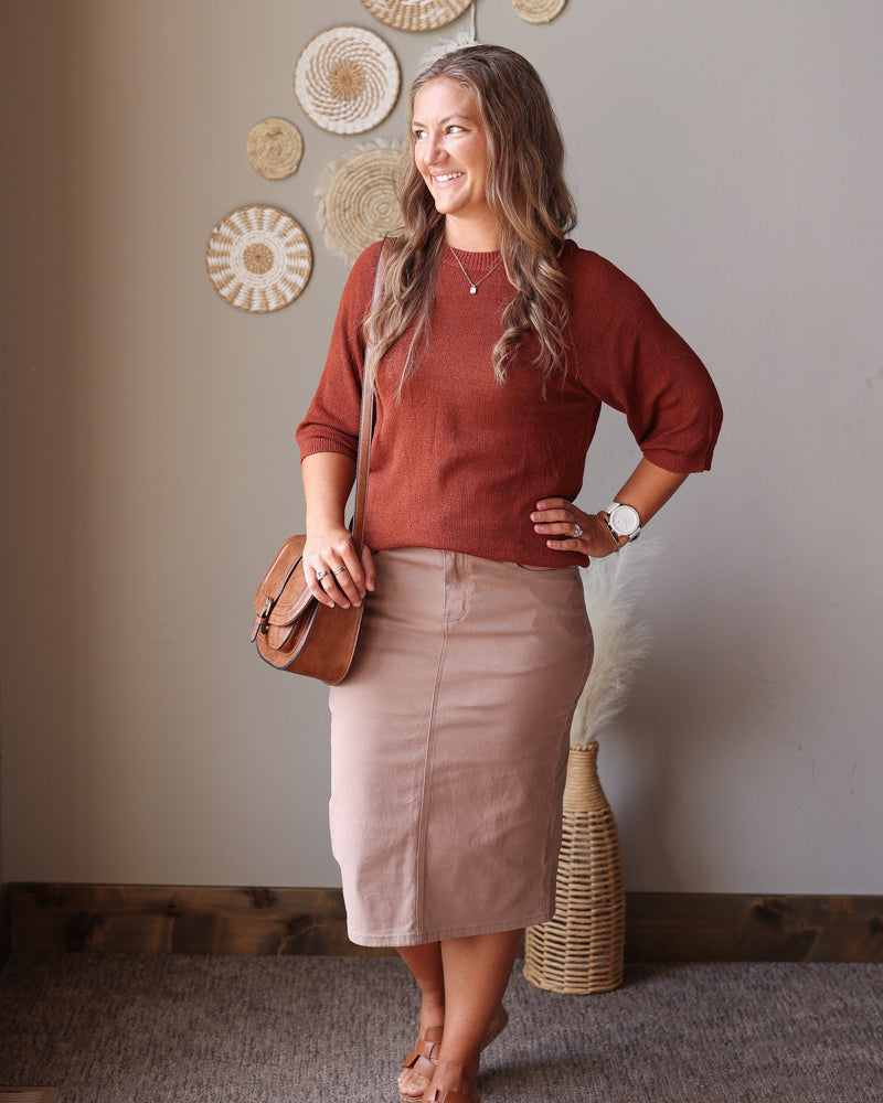 A woman modeling a rust-colored long-sleeve knit top and a khaki taupe pencil skirt, carrying a brown leather crossbody bag. She is standing in a room with a neutral beige wall decorated with circular woven wall art.