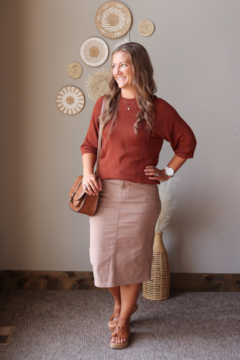 A woman modeling a rust-colored long-sleeve knit top and a khaki taupe pencil skirt, carrying a brown leather crossbody bag. She is standing in a room with a neutral beige wall decorated with circular woven wall art.