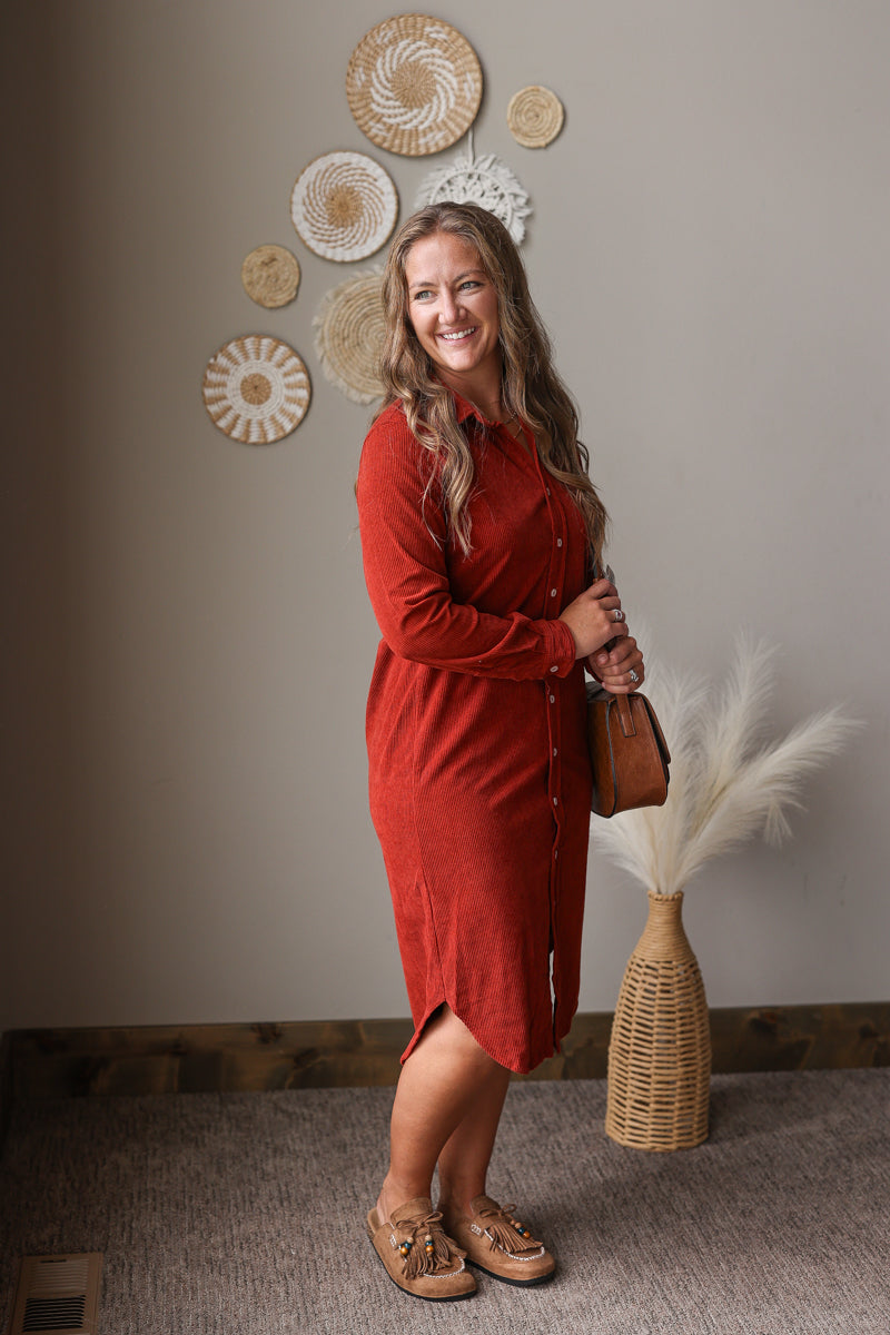 A woman wearing a rust-orange long-sleeve button-up shirt dress, holding a brown leather bag and wearing tan loafers, standing in a styled room with boho decor.