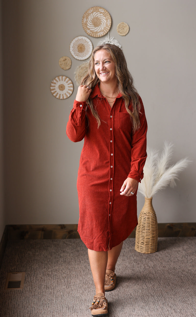 A woman wearing a rust-red corduroy button-down shirt dress, standing in a room with a neutral gray wall decorated with circular wall art and a woven vase with pampas grass.