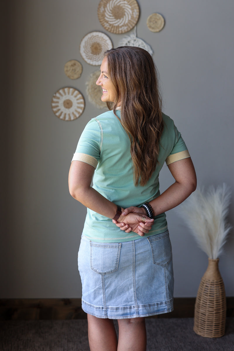 A woman with long brown hair is shown from behind wearing a mint green short-sleeve t-shirt with beige stripes on the sleeves and a light blue denim skirt. She is standing in a room with a gray wall decorated with circular woven wall art.