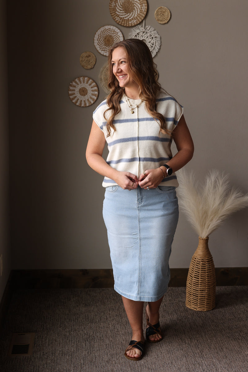 Woman wearing a striped shirt and denim skirt standing in a room with decorative wall art and a vase.