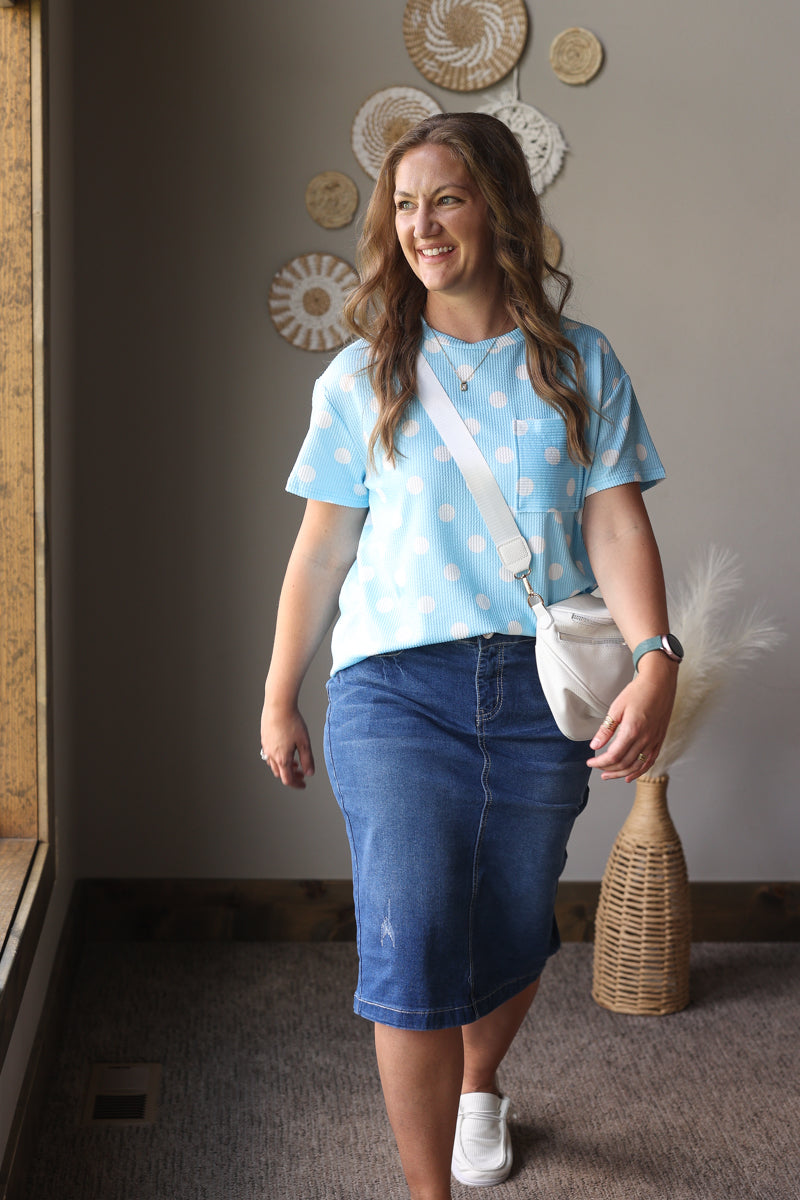 Woman wearing a light blue polka dot shirt and denim skirt standing indoors.