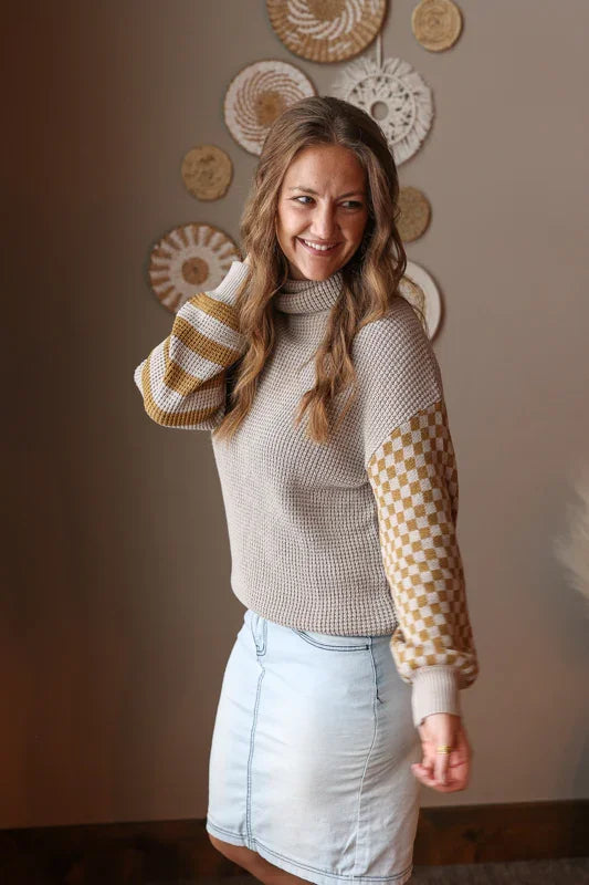 Woman wearing a grey turtleneck sweater with checkered sleeves and a light blue denim skirt, standing against a wall with decorative plates.