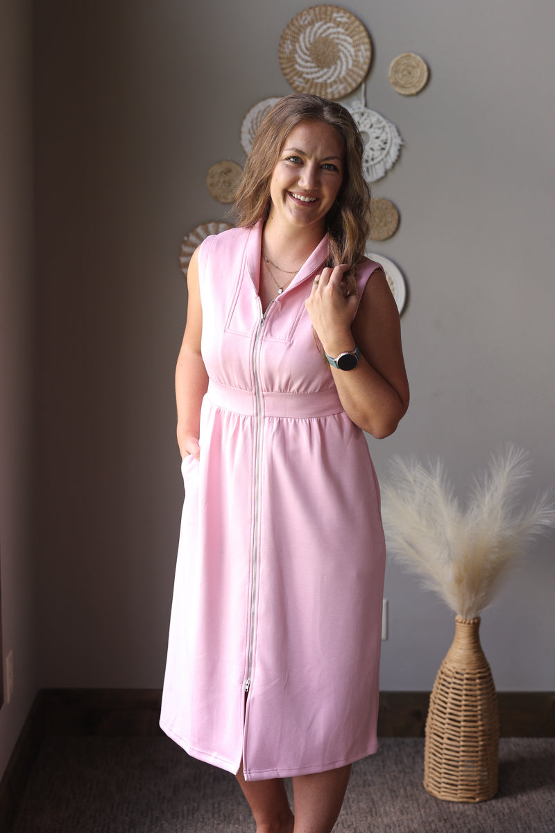 Woman wearing a pink dress standing in a room with decorative wall art and a vase.