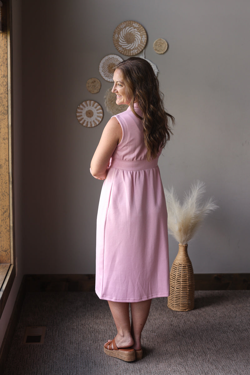 Woman in a pink dress standing in a room with decorative wall art and a vase.