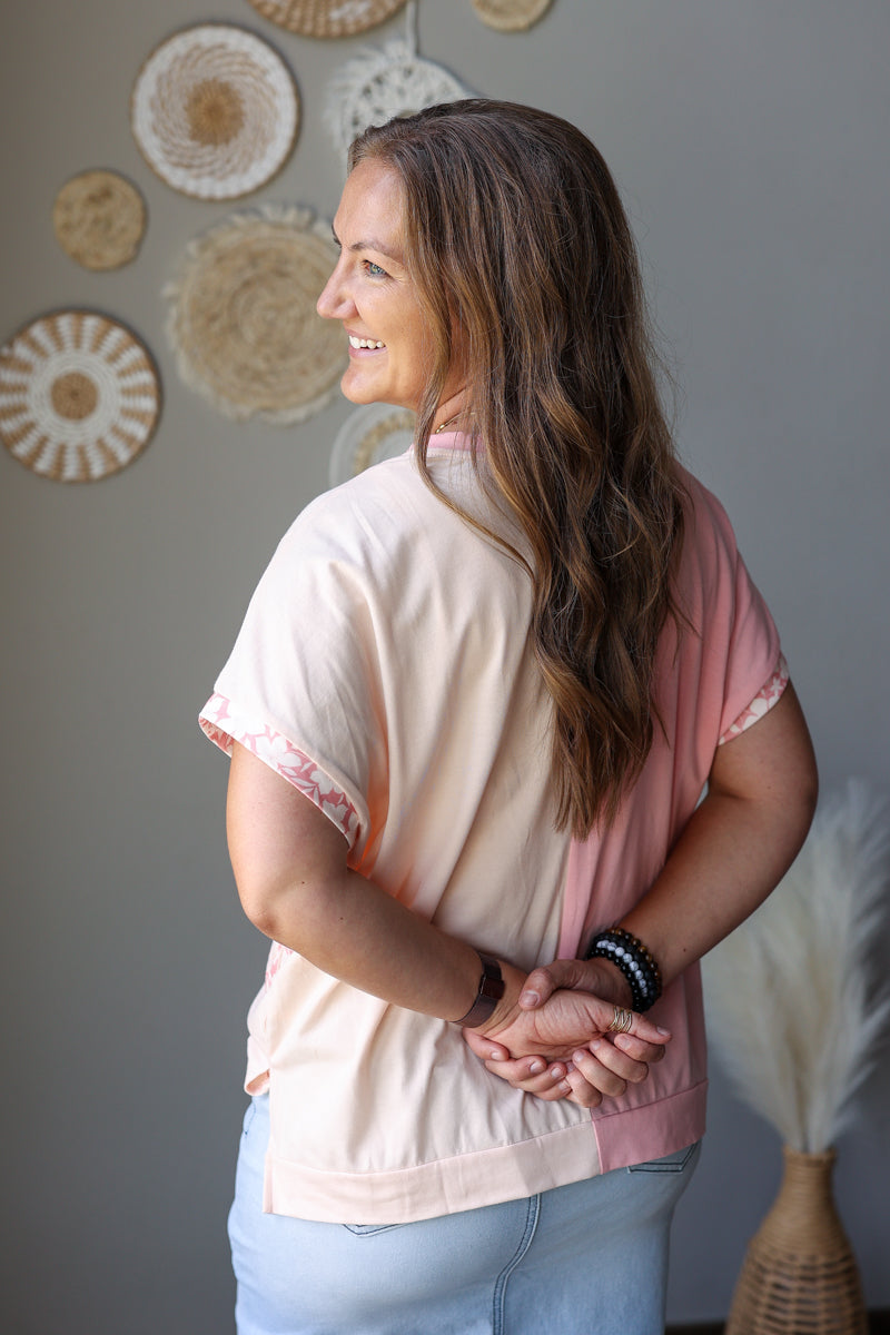 A woman with long brown hair is shown from the back and side, wearing a short-sleeve top with a split design of light beige and dusty pink, paired with light blue jean skirt. She is standing in a room with a gray wall decorated with circular woven wall art.