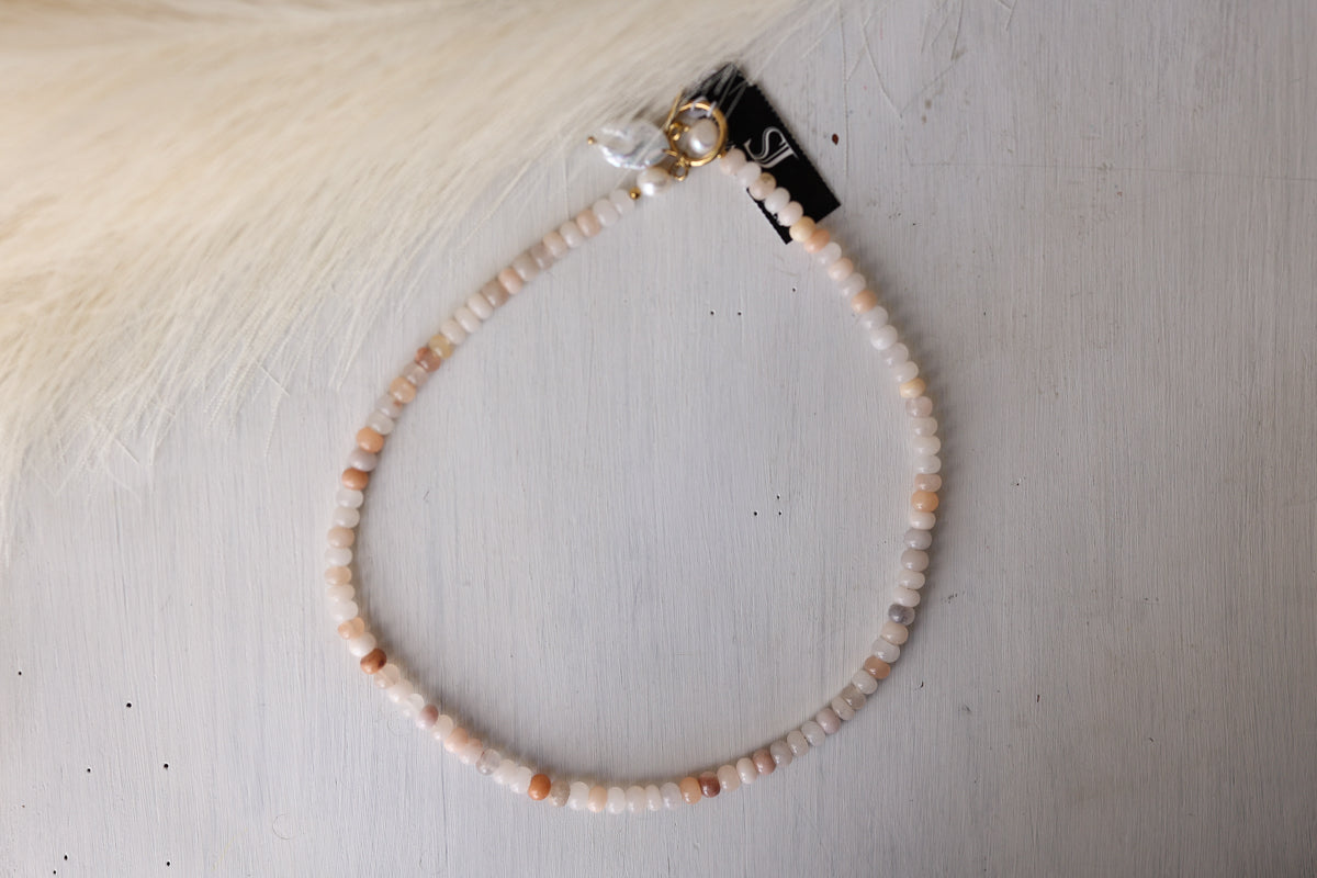 A beaded necklace made of small peach, cream, and white stone beads with a gold-toned clasp, laid on a light gray surface with a cream faux-fur prop at the top left.