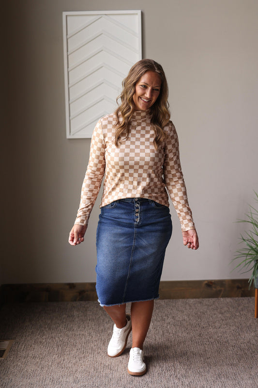 Woman wearing a checkered top and denim skirt standing in a room with a neutral wall and plant for fall outfits.