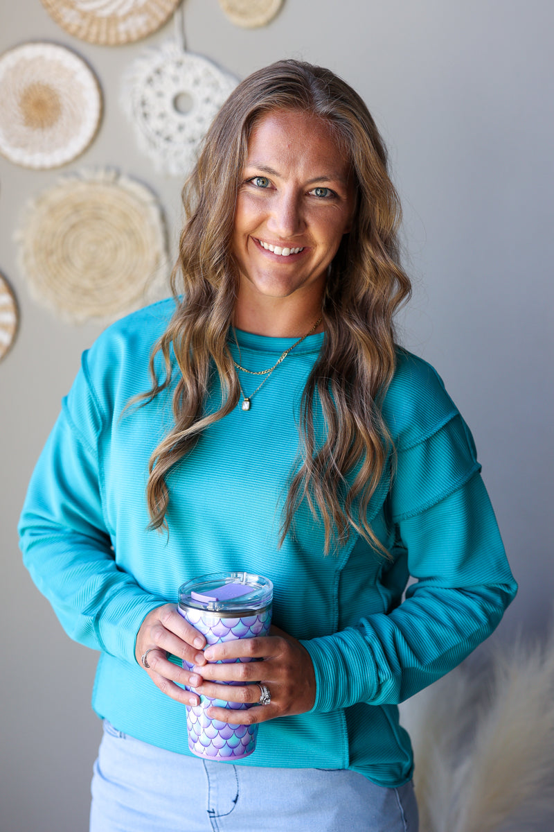 A smiling woman with long wavy brown hair wearing a bright turquoise long-sleeve top and light blue jean skirt, holding a purple and white mermaid-scale patterned tumbler cup. She is standing indoors against a gray wall decorated with circular woven wall hangings.