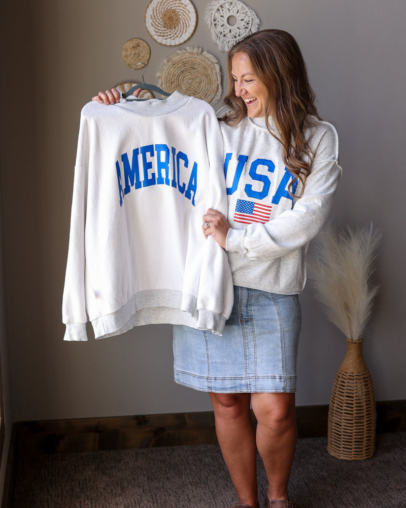 Woman holding a white sweatshirt with 'AMERICA' and 'USA' text, standing in a room with decorative items on the wall.