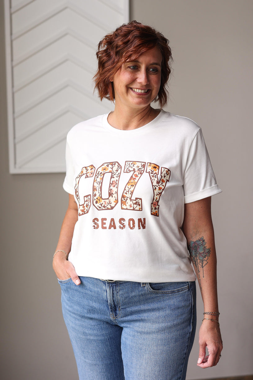 Person wearing a white t-shirt with 'Cozy Season' text and floral design, standing indoors.