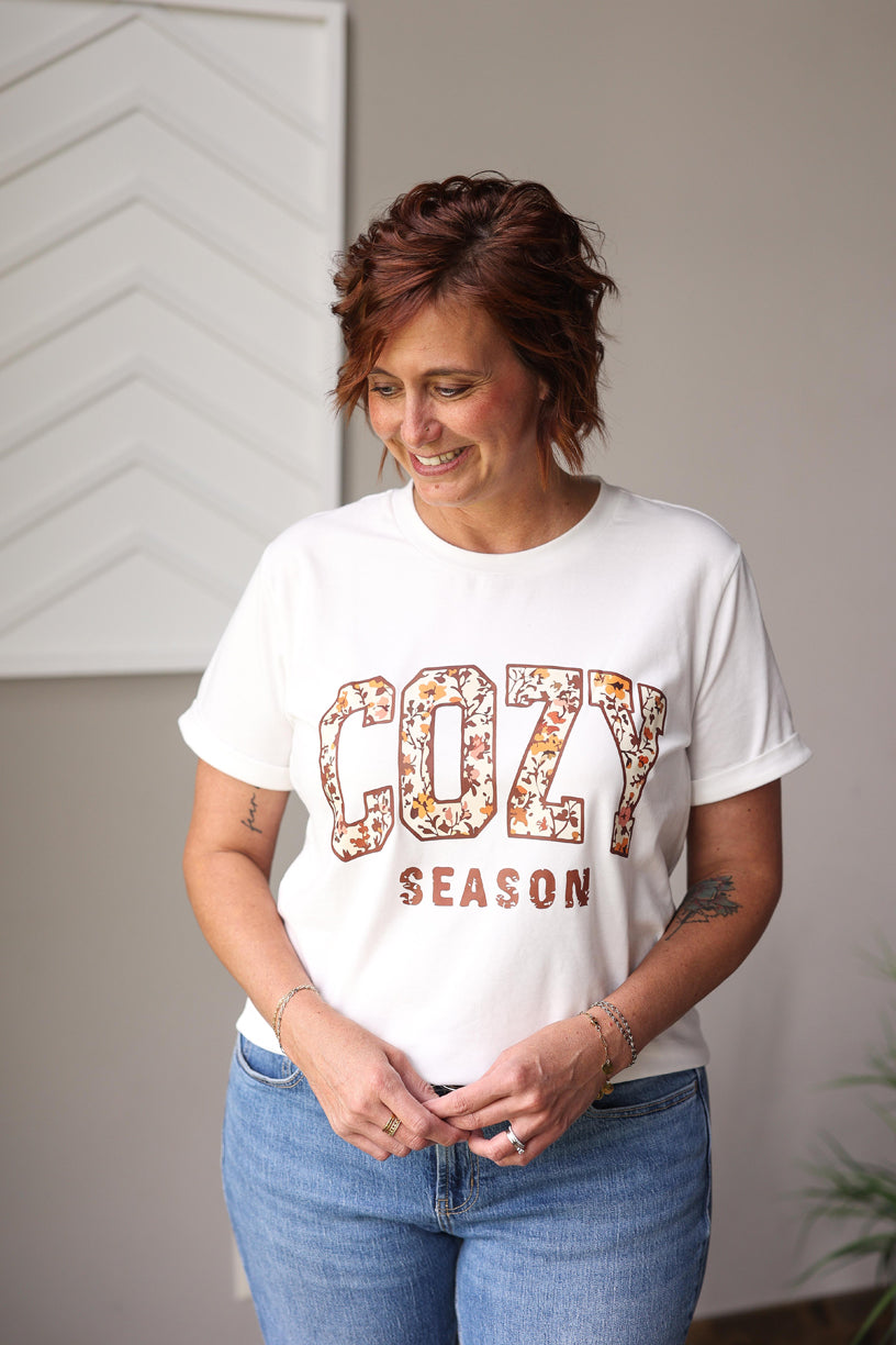 Woman wearing a white t-shirt with 'COZY SEASON' text and floral design, standing indoors.