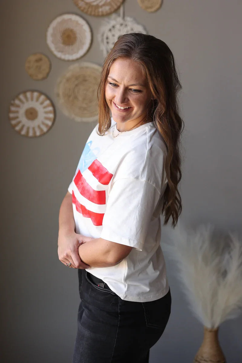 Woman wearing a white t-shirt with a red graphic design, standing against a gray wall with decorative items.