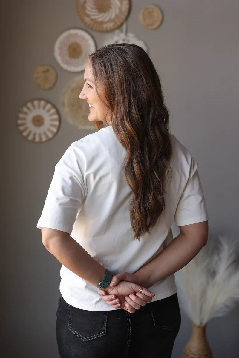 Woman in a white shirt standing with her hands clasped in front of a wall with decorative circular elements.