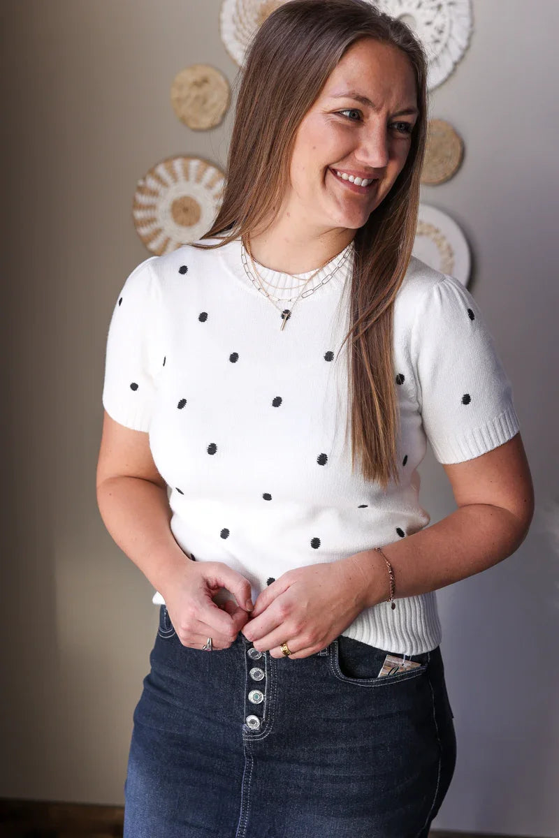 Woman wearing a white polka dot shirt and dark jeans with decorative plates in the background.