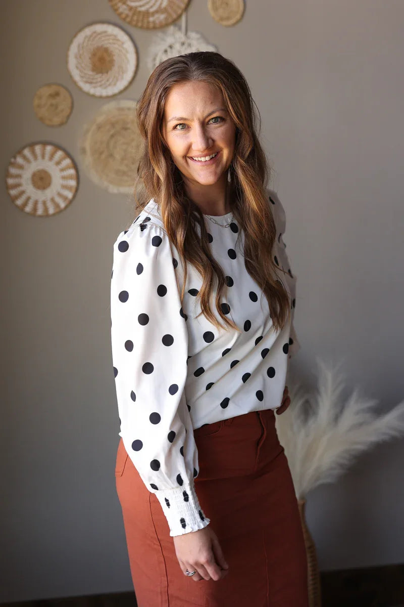 Woman wearing a polka dot blouse and rust midi skirt standing against a neutral wall with decorative elements.
