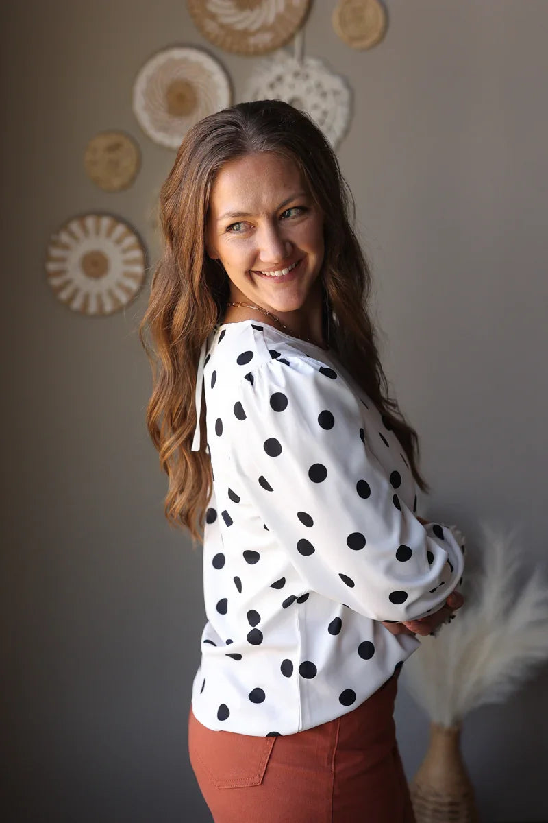Woman wearing a white blouse with black polka dots and rust denim skirt, standing against a neutral wall with decorative elements.