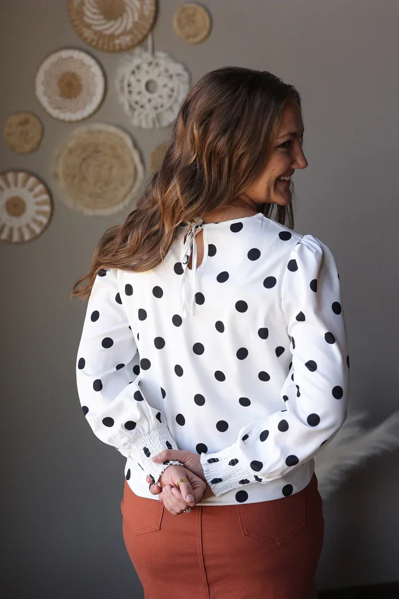 Woman wearing a white blouse with black polka dots and rust midi skirt, standing against a wall with decorative circular patterns.