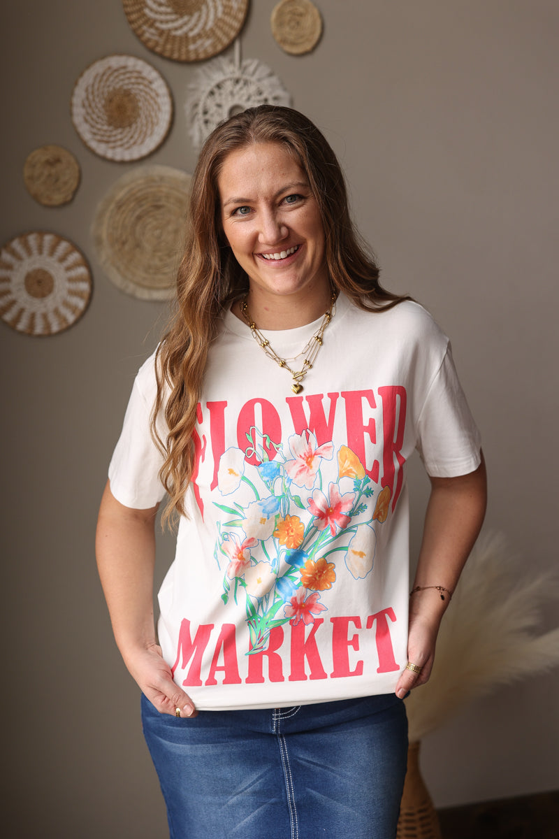 Woman wearing a white t-shirt with floral design and text, standing against a wall with decorative items.