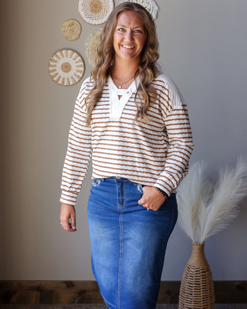 A woman modeling a cream and brown horizontally striped long-sleeve knit top with a white V-neckline, paired with a blue denim skirt, standing in front of a gray wall with decorative woven wall art.