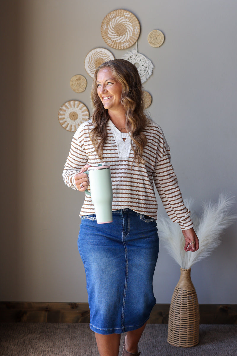 A woman wearing a beige and brown striped long-sleeve top and a blue denim skirt, holding a mint green tumbler. She is standing in a room with a gray wall decorated with circular woven wall art, a wicker basket, and a vase with pampas grass.