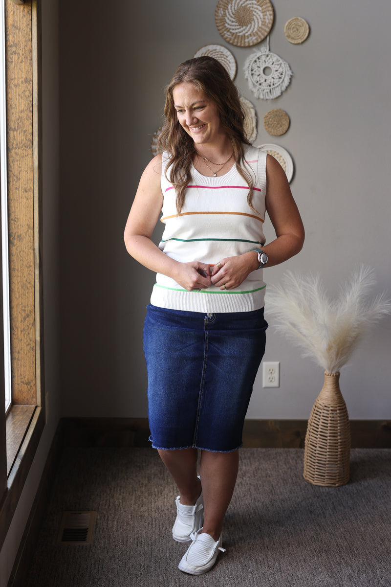 Woman standing in a room with decorative wall art and a vase wearing a white striped sweater tank top and denim skirt.