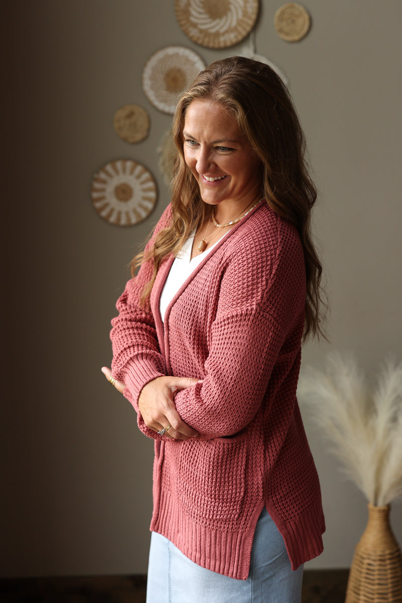 A woman wearing a dusty rose waffle-knit cardigan over a white top and light blue jean skirt, standing in a warmly lit room with decorative wall art and pampas grass.