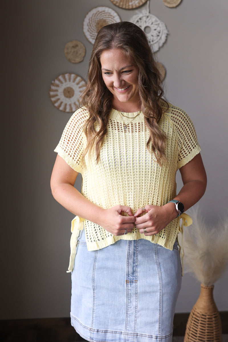 Woman wearing a yellow crochet top and blue skirt standing in a room with decorative wall hangings.