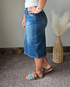 A person wearing a blue denim midi skirt, styled with a white top and brown slide sandals, standing in an indoor setting with a woven vase of pampas grass in the background.