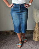 A person wearing a medium-wash blue denim midi skirt with a front fly, paired with a white top and blue sandals, standing indoors next to a woven vase with dried grass.