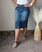 A person wearing a knee-length blue denim skirt with a side slit, paired with a white top and brown slide sandals, standing indoors against a gray wall with a woven vase of white pampas grass to the right.