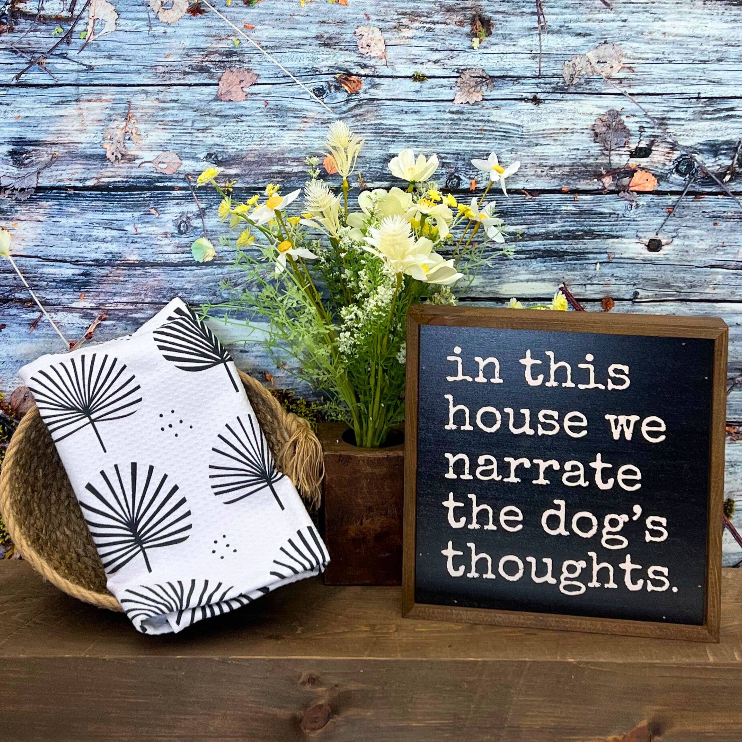 A styled home decor scene on a rustic wooden surface with a blue distressed wood plank backdrop. On the left, a folded white cloth napkin or towel with a black palm frond pattern sits in a woven basket.