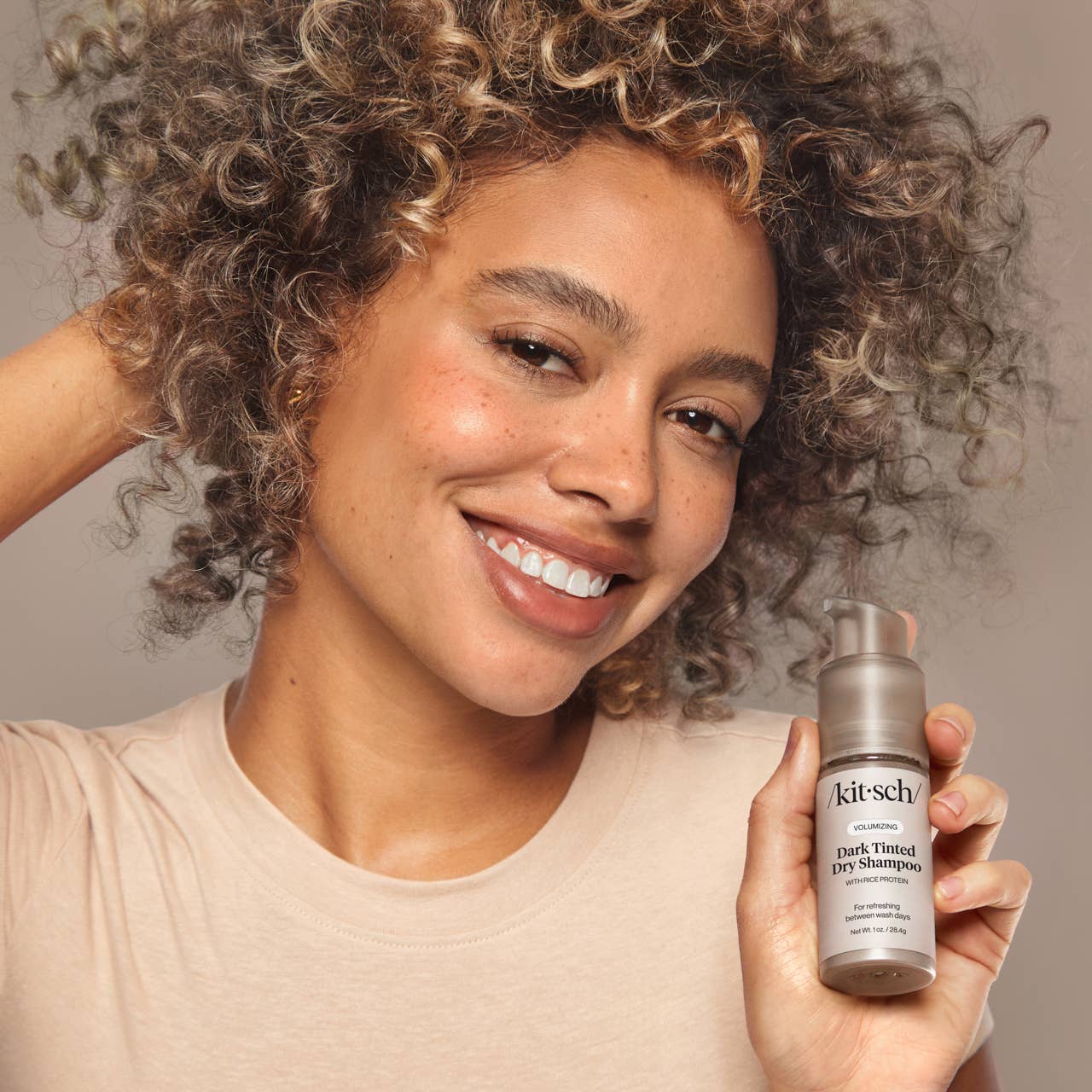 Woman holding a bottle of 'kitsch' dark tinted dry shampoo against a neutral background