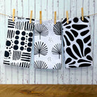 Three white kitchen towels with black abstract and botanical patterns hang from a string on wooden clothespins against a whitewashed wood plank wall.