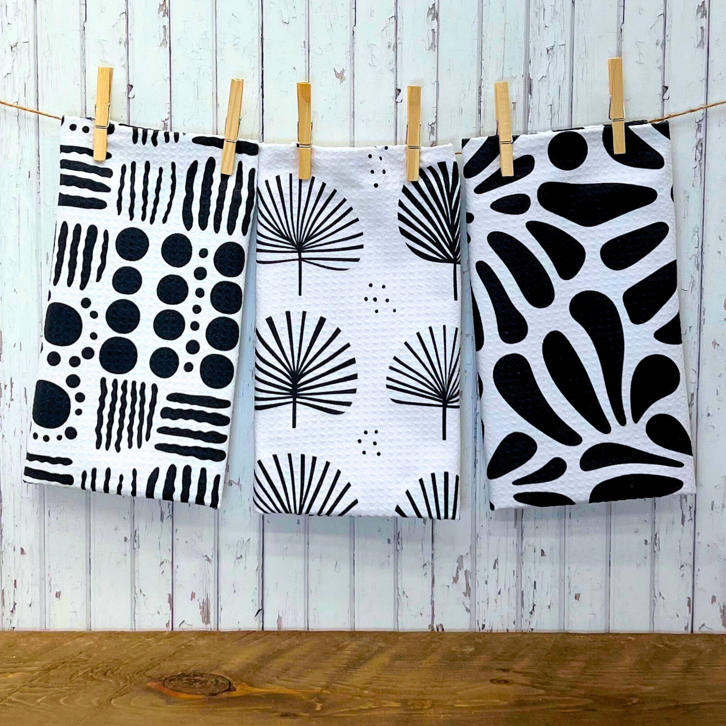 Three white kitchen towels with black abstract and botanical patterns hang from a string on wooden clothespins against a whitewashed wood plank wall.