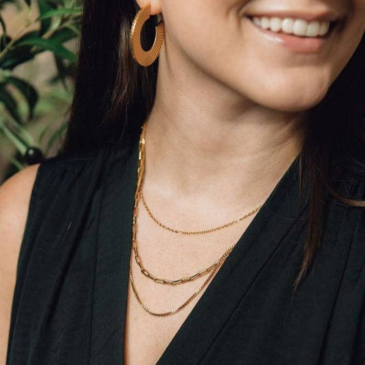 Woman wearing gold hoop earrings and layered gold necklaces with a blurred green plant background